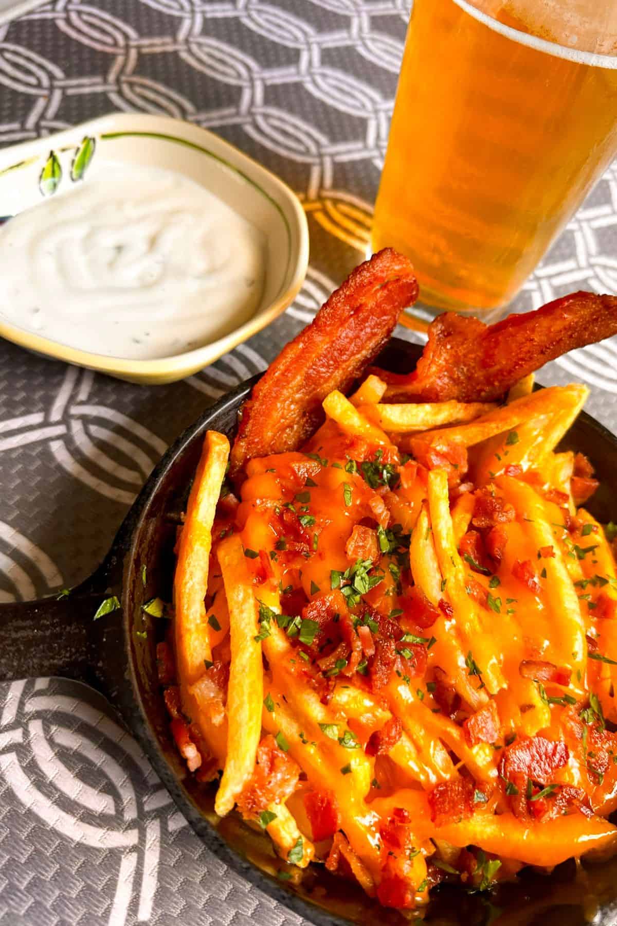 Small cast iron pan full of bacon cheese fries with a cold beer and small bowl of ranch